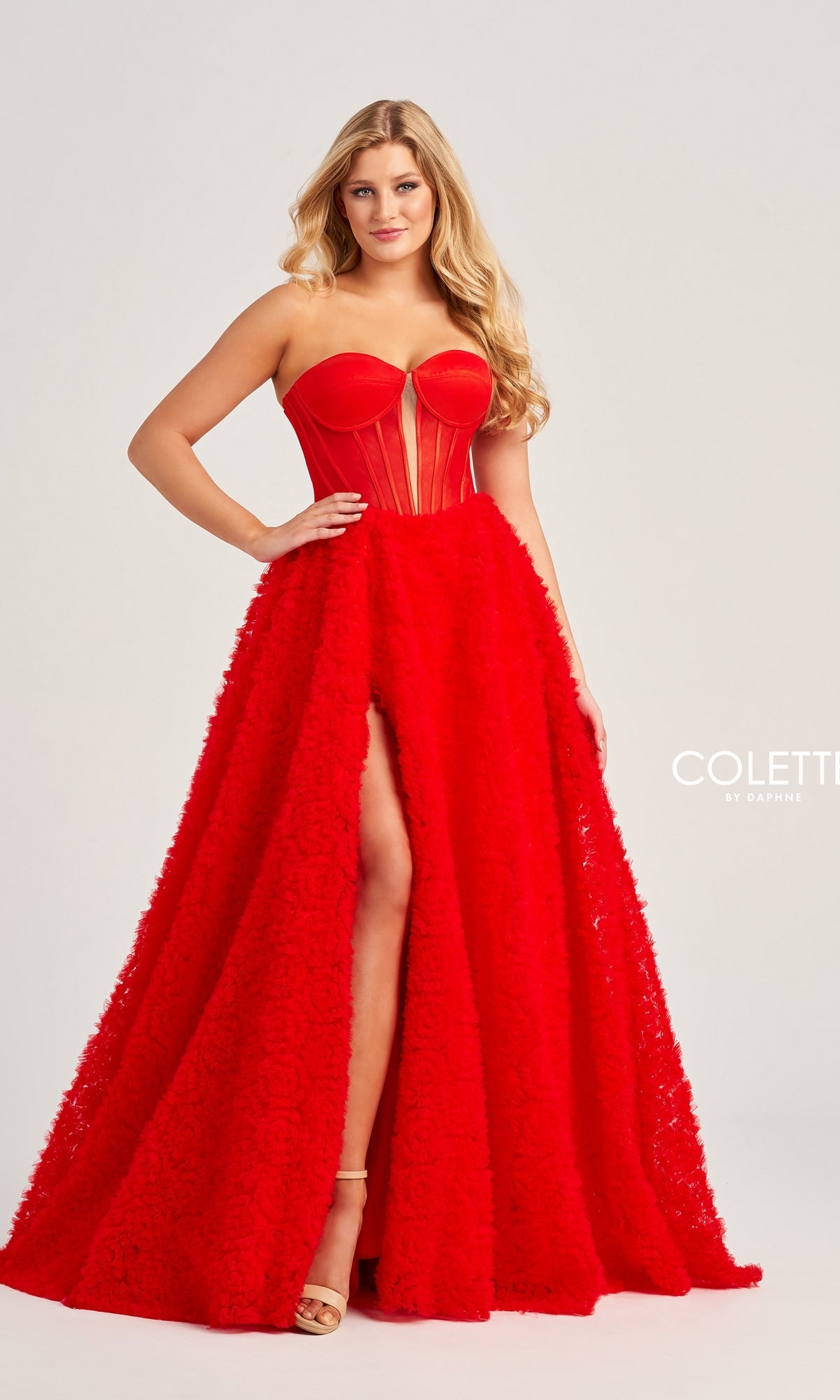 A woman poses elegantly in the Bustier Colette Long A-Line Prom Ball Gown CL5114, a strapless bright red dress by Colette by Daphne featuring a fitted bodice, sheer paneling, and a high skirt slit against a plain background.